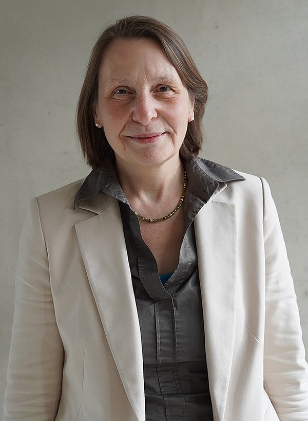 Prof. Dr. Elke Theobald in grauer Bluse und weißem Blazer vor grauem Hintergrund