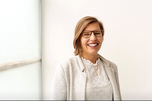 Dominique Schuppler is wearing glasses, a white blouse, and a blazer. She is smiling at the camera in a light-filled room.