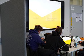 Zwei Studierende arbeiten an Laptops, im Hintergrund ein großer Bildschirm, der eine Präsentation zeigt