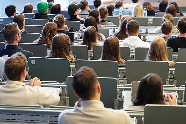 Students are sitting in a full lecture hall with notes on their desks