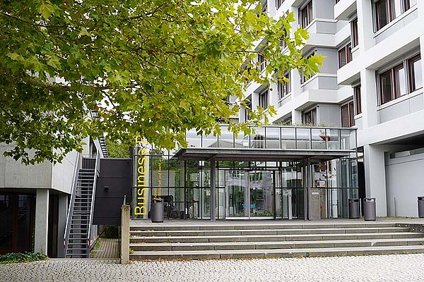 Der Haupteingang der Business School Pforzheim im Sommer mit einem in grün leuchtenden Laubbaum davor