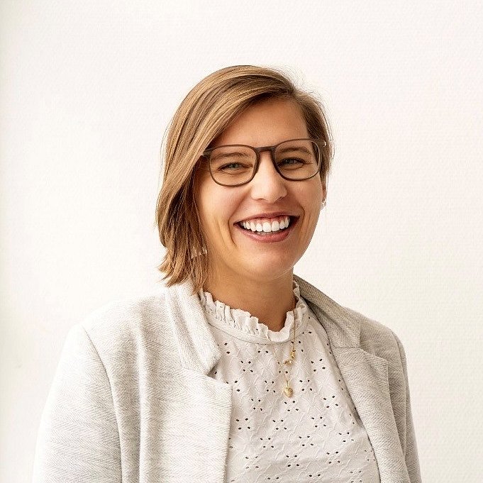 Dominique Schuppler is wearing glasses, a white blouse, and a blazer. She is smiling at the camera in a light-filled room.