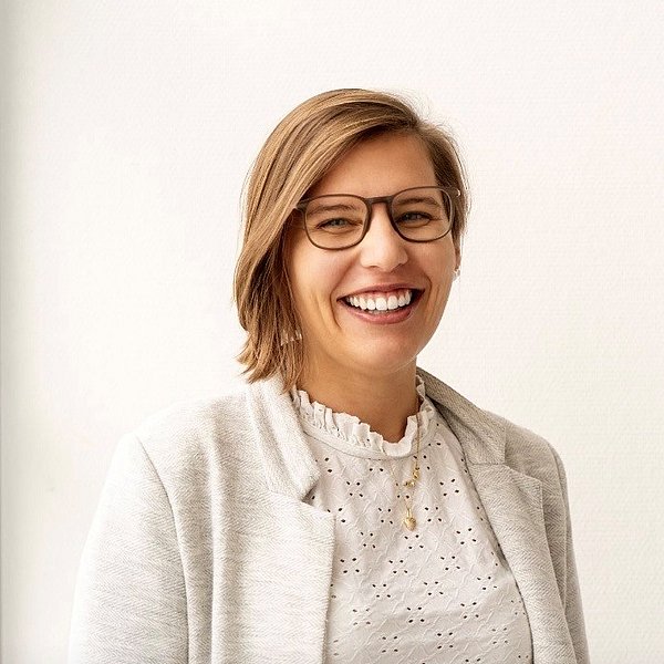 Dominique Schuppler is wearing glasses, a white blouse, and a blazer. She is smiling at the camera in a light-filled room.