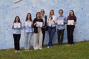 Prof. Dr. Sabine Assmann smiles at the camera with her students, who have received a Beta Gamma Sigma award, in front of a blue wall