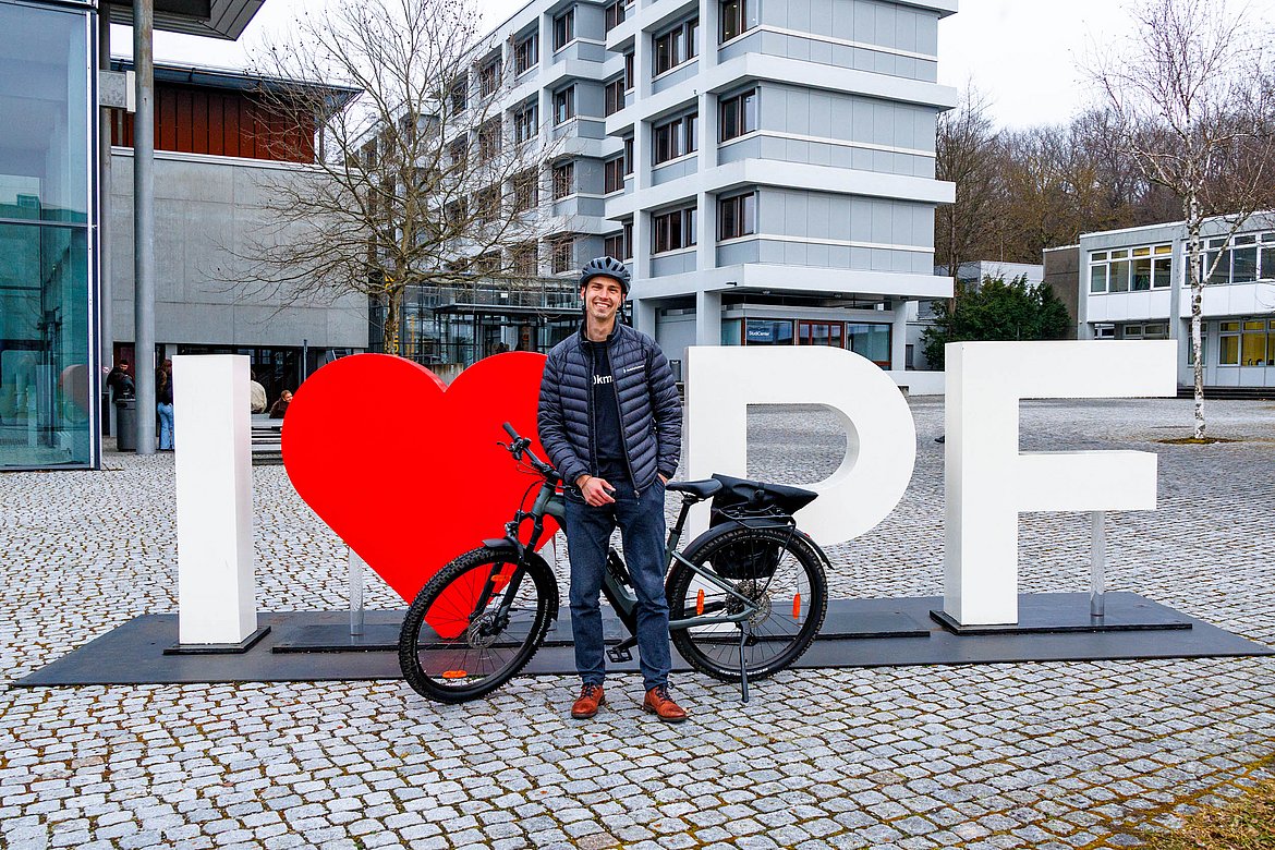 John steht mit einem Fahrrad vor der Hochschule und lächelt in die Kamera.