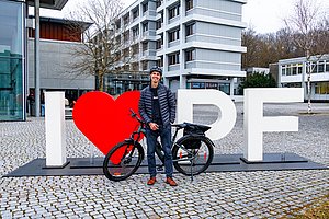 John steht mit einem Fahrrad vor der Hochschule und lächelt in die Kamera.