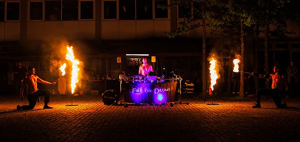 Zwei Männer mit Fackeln machen bei der Feuershow eine Choreo, während ein dritter an einer Trommel für musikalische Untermalung sorgt.