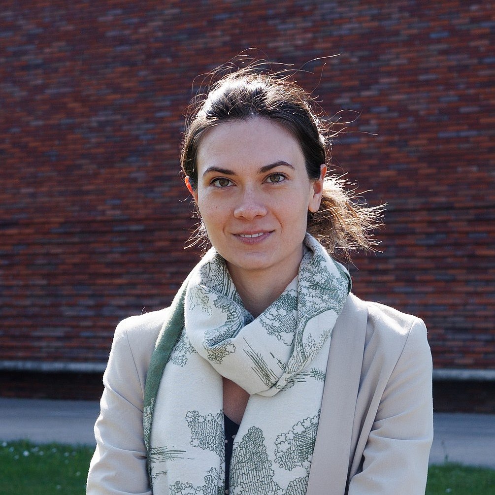 Ms. Bidler is sitting on campus and smiling at the camera. She is wearing a blazer and has her brown hair tied back. 