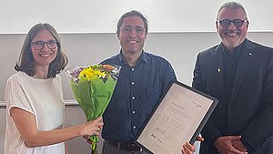 Award recipient Prof. Dr. Fernando Fastoso holding a certificate and a bouquet of flowers, with Prof. Dr. Anja Spilski standing to his left and Prof. Dr. Thomas Cleff standing to his right
