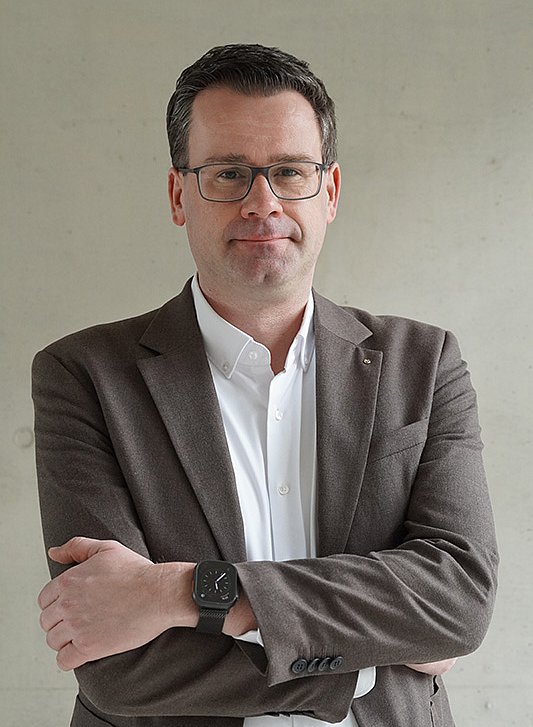 Prof. Dr. Torben Kuhlenkasper mit braun meliertem Sakko und weißem Hemd vor grauem Hintergrund