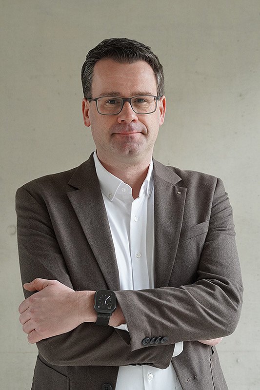 Prof. Dr. Torben Kuhlenkasper mit braun meliertem Sakko und weißem Hemd vor grauem Hintergrund