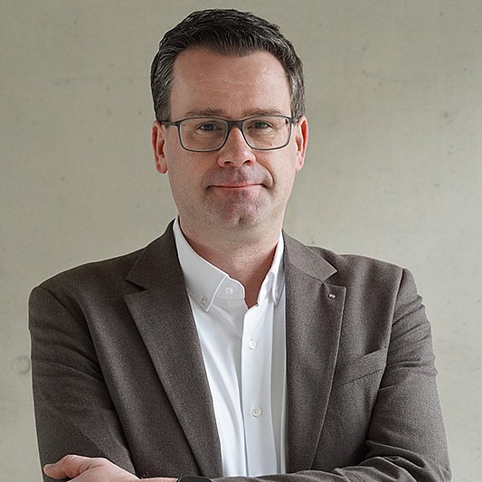 Prof. Dr. Torben Kuhlenkasper mit braun meliertem Sakko und weißem Hemd vor grauem Hintergrund
